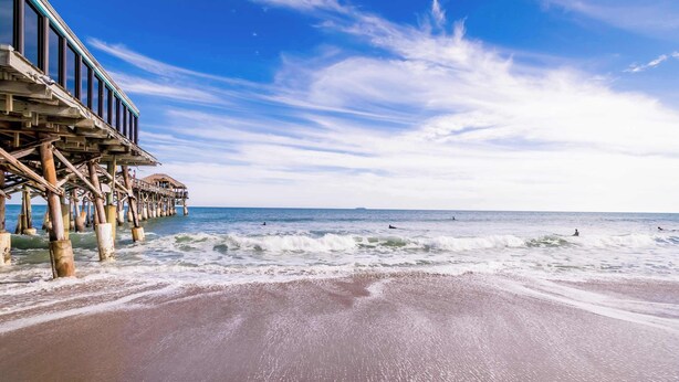 Una playa con un muelle de madera que se adentra en el océano, olas rompiendo en la orilla y un cielo despejado.