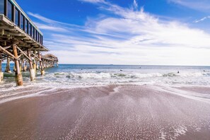 Une plage avec une jetée en bois qui s’avance dans l’océan, des vagues qui se brisent sur le rivage et un ciel dégagé.