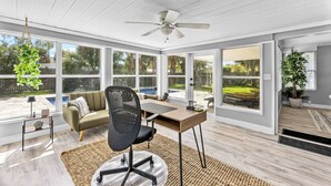 Un solárium con un escritorio de madera, una silla de oficina negra, un sofá y ventanas amplias con vista a un patio trasero.