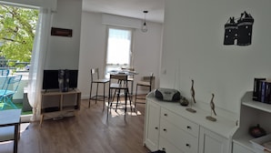 A modern living room with a dining area, wooden flooring, a white cabinet with decorative items, and a large window providing natural light. - Apartment in the center of Guérande 4 minutes walk from the ramparts (Guérande)