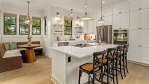 A modern kitchen with white cabinetry, a large island, bar stools, and a dining area with a bench. - Hale Kohola by Alekona Kauai: Brand New 4BR Home w AC, close to beaches, shopping, dining (Koloa)