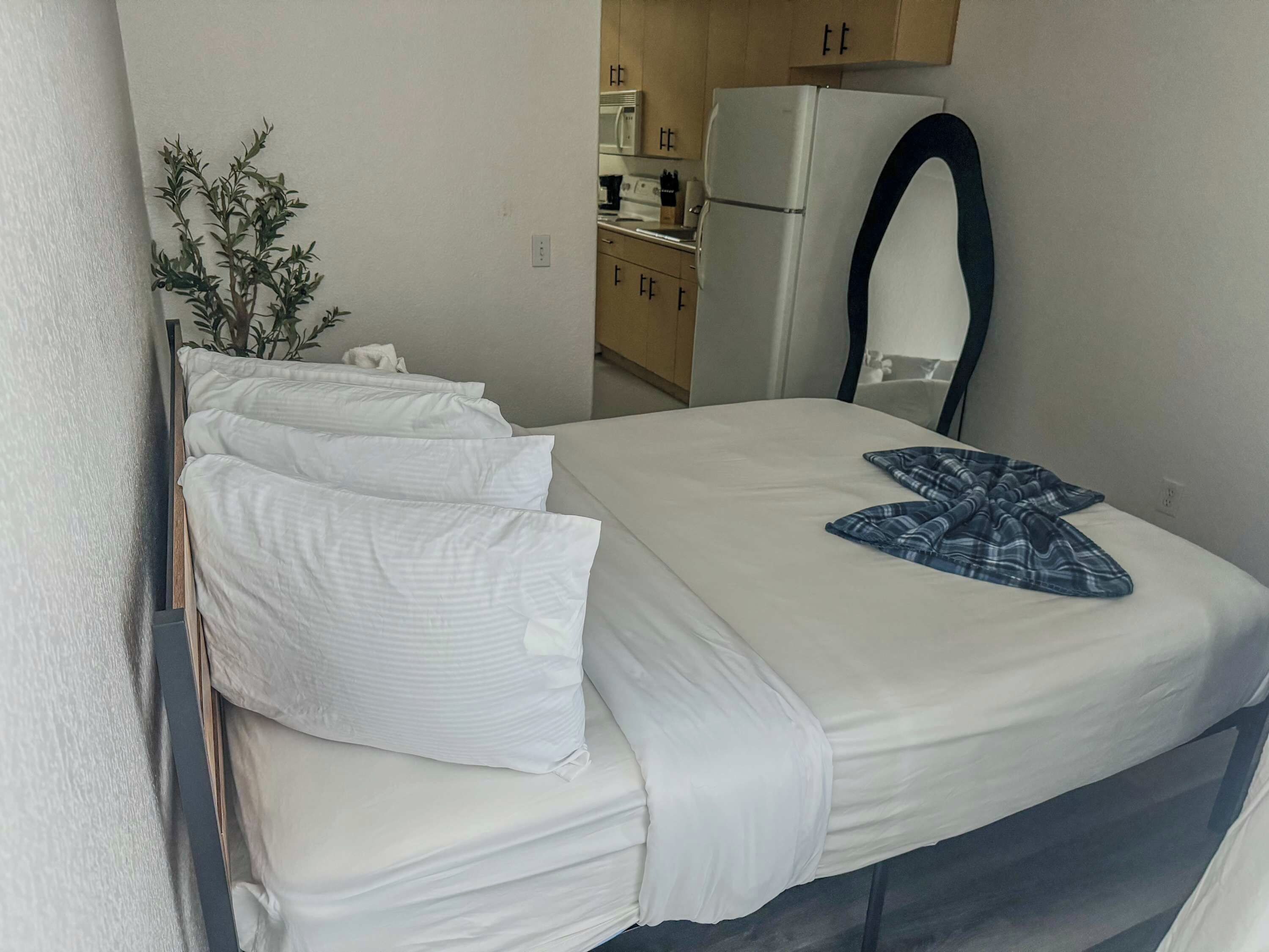 Ein ordentlich bezogenes Bett mit weißen Leinen und einem blauen Kopfkissen, eine kleine Topfpflanze und ein weißer Kühlschrank in einem kleinen Zimmer.