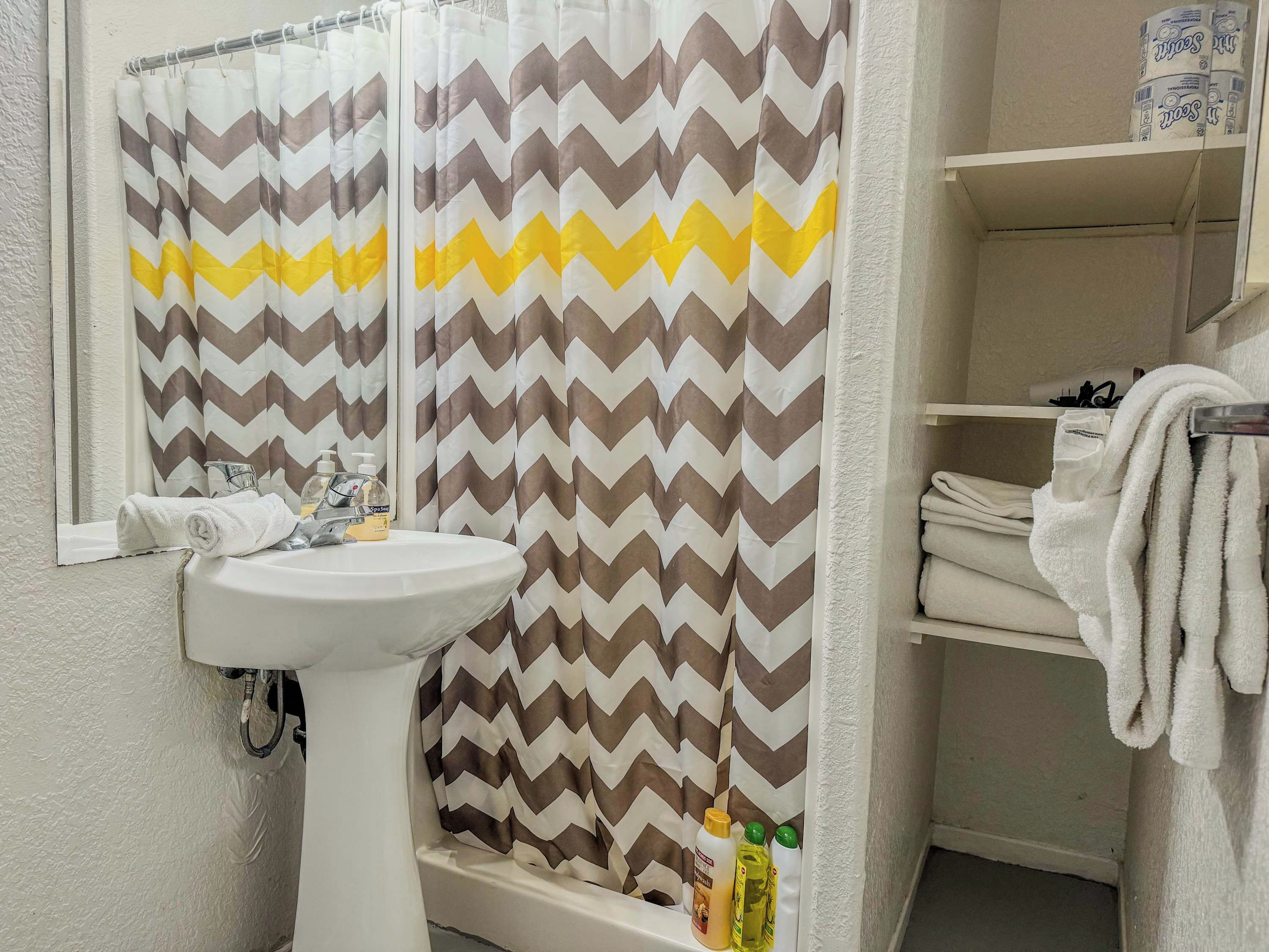 A bathroom with a white sink, a mirror, a shower curtain with a zigzag pattern, and a shelf with towels.