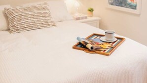 A neatly made bed with a white comforter, a beaded pillow, and a tray with a book and a cup on the bedside table. - La Jolla Suite with Ocean View Patio (San Diego)