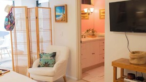 A room with a white armchair, a television mounted on the wall, a wooden side table, and a kitchen area visible through an open door. - La Jolla Suite with Ocean View Patio (San Diego)