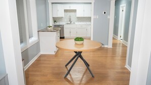 Cozinha moderna com armários brancos, uma mesa de jantar de madeira com uma planta, e um corredor que leva a uma porta.