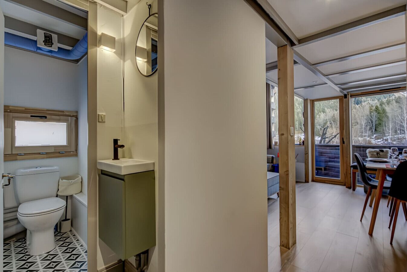 Ein kleines Badezimmer mit Toilette, Waschbecken und Spiegel. Es hat ein Fenster mit Jalousien und Blick auf eine bewaldete Landschaft.