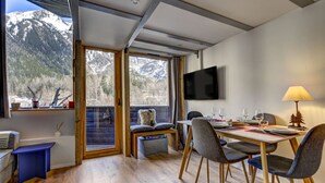 Un espace de repas moderne avec une table dressée pour deux personnes, un téléviseur fixé au mur et une vue sur des montagnes enneigées, visible à travers de grandes fenêtres.