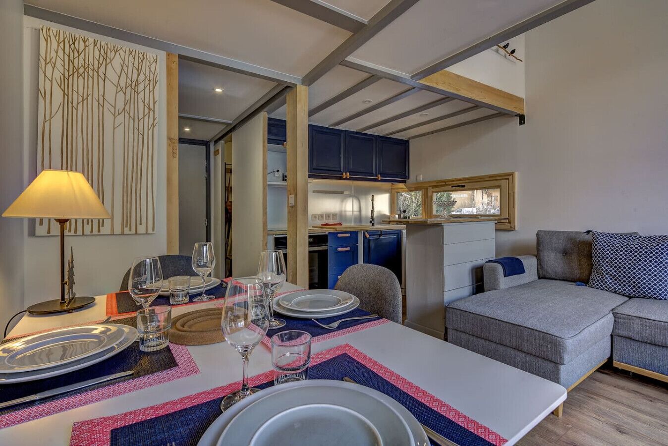 Un comedor con una mesa dispuesta para cuatro personas, un sofá y una zona de cocina con armarios y un ventanal.