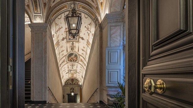 Un vaste hall d’entrée avec un sol à carreaux, une grande porte richement décorée et un escalier menant à un palier doté d’une cheminée.