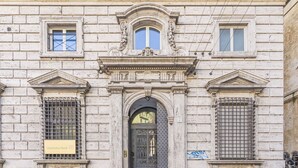 Une façade d’immeuble avec une entrée centrale, flanquée de deux fenêtres dotées de grilles en métal, et une plaque portant l’inscription « Deutsche Bank ».