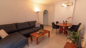A living room with a dark grey sofa, a wooden coffee table with a candle and a plant, and a dining area with a round table and chairs.