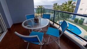 A balcony with a glass table, blue chairs, and a view of the ocean and palm trees.