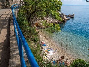 Eine Küstenstraße mit blauer Lehnschranke, klarem Wasser und einem Strand mit badenden Personen.