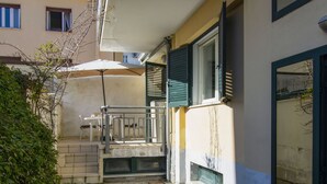 Uma pequena área externa com uma mesa, cadeiras, um guarda-sol branco e uma escada que leva a uma porta.