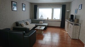 Un salon avec un canapé gris, une petite table et un meuble en bois. Il y a deux fenêtres avec des rideaux, un radiateur et une lampe sur une table.
