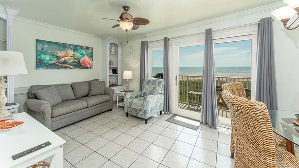 A living room with a grey sofa, a patterned armchair, a ceiling fan, and a view of the ocean through large sliding glass doors.