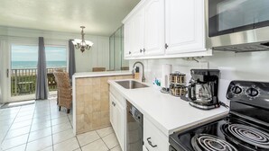 A kitchen with white cabinets, a stove, a sink, and a coffee maker. There is a balcony with a view of the ocean.