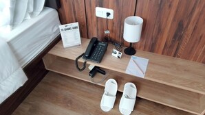 Une chambre d’hôtel avec un lit en bois, une table de chevet avec un guide des programmes télévisés, un téléphone, une lampe, des chaussons et un bloc-notes.
