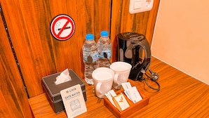 Un espace aménagé dans une chambre d’hôtel, comprenant un panneau « défense de fumer », des bouteilles d’eau, une machine à café, des tasses, des mouchoirs et un panneau « protégeons l’environnement ».
