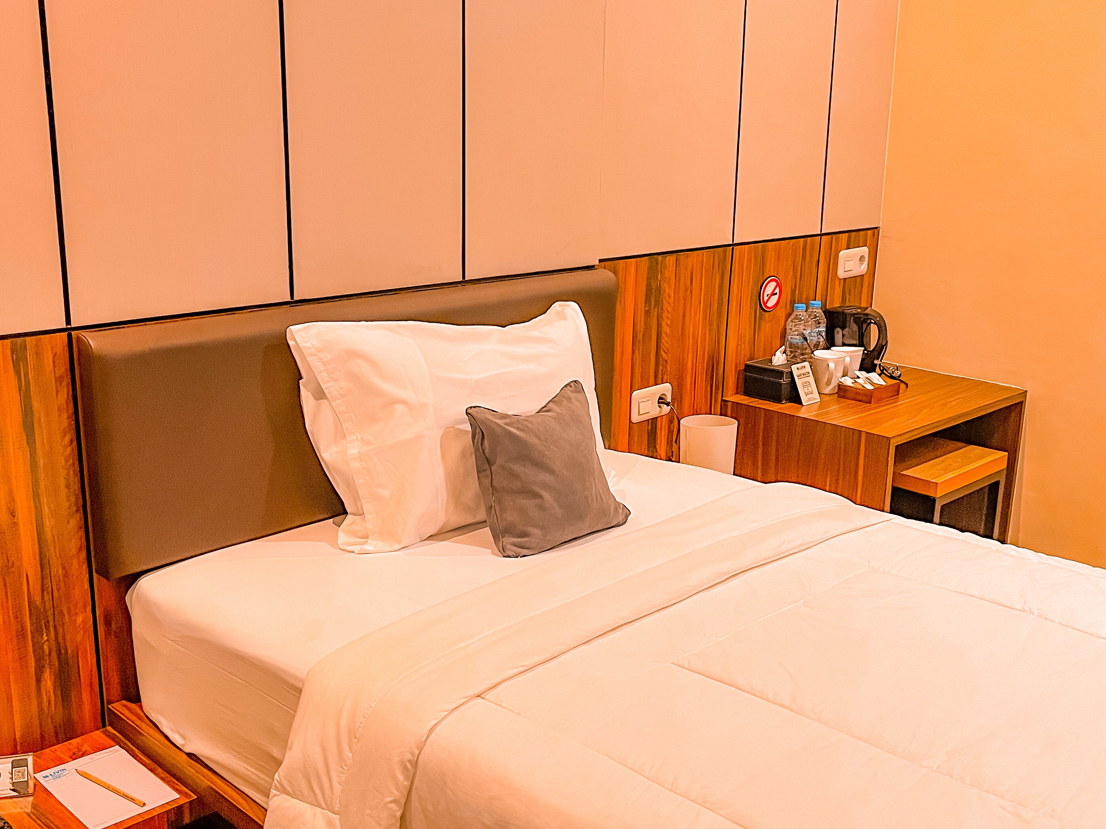 A hotel room with a bed, pillows, a wooden nightstand, a coffee maker, and a sign indicating no smoking.