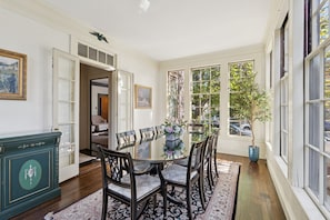 A dining room with a round table, chairs, and a large window with a view of trees and parked cars.