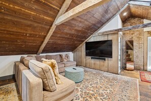 An attic room with a wooden ceiling, a large TV mounted on the wall, a sofa, and a patterned rug.