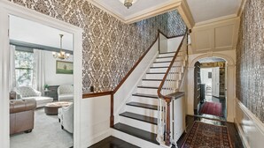 A staircase with a wooden handrail and black steps, leading to a room with patterned wallpaper and a chandelier. - Charming Historic Estate | In-town location (Portland)