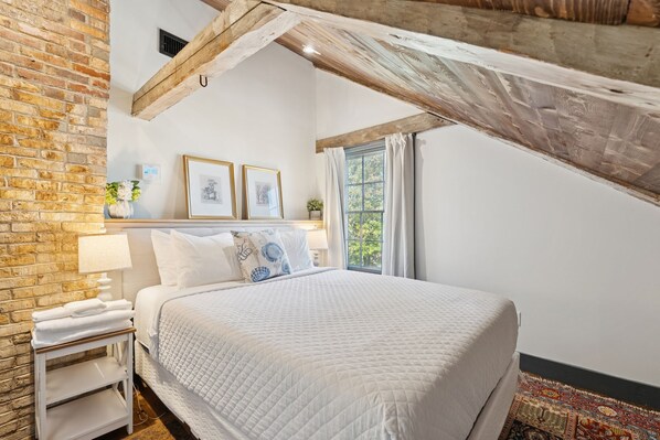A bedroom with a brick wall, a wooden ceiling beam, a bed with white bedding, a nightstand with a lamp, and a window with curtains.