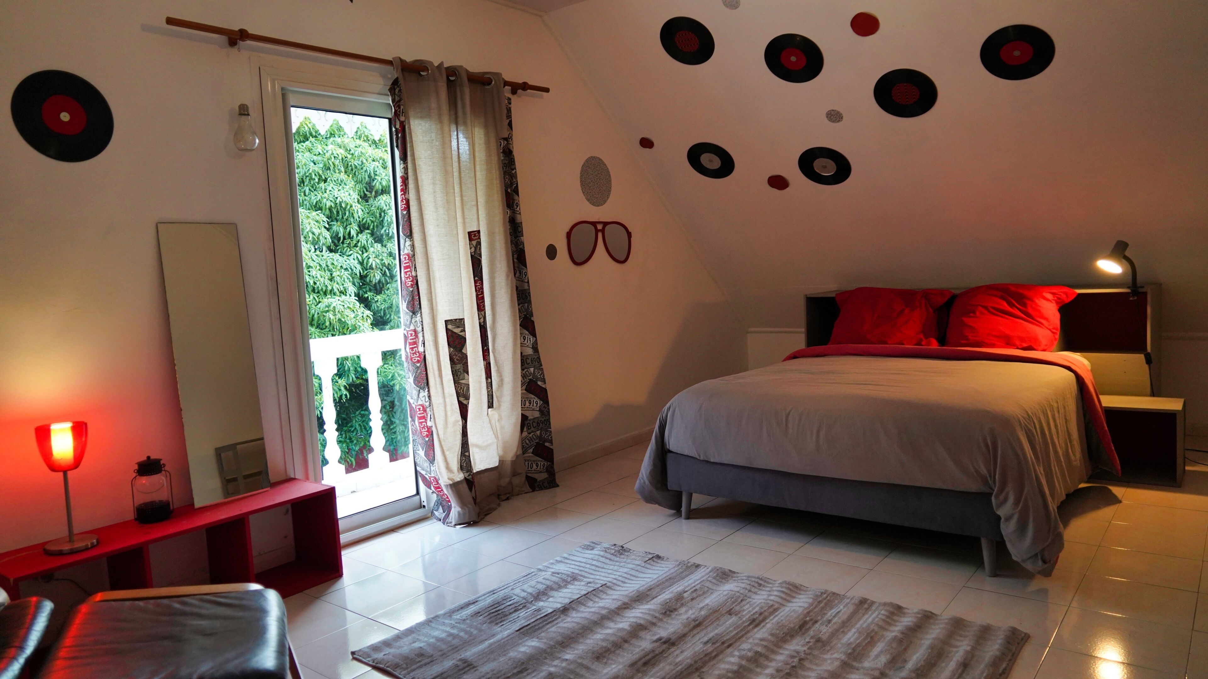 Une chambre à coucher avec un lit, des oreillers rouges, une table de chevet, une lampe, une fenêtre avec des rideaux et des éléments de décoration murale, notamment des disques vinyle.