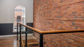 Een trap met een houten leuning en zwarte metalen spijbels, die leidt naar een kamer met een bakstenen muur en een raam met uitzicht op een ander bakstenen gebouw.