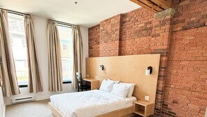 Een moderne hotelkamer met een bakstenen muur, een houten plafond, een groot bed, een bureau en een stoel.