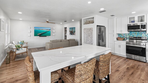 Una cocina moderna con muebles blancos, una isla grande con sillas de mimbre, una mesa de comedor y un televisor montado en la pared.