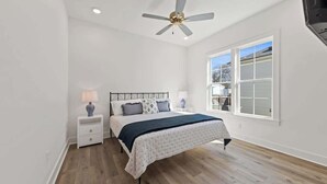 Une chambre à coucher comprenant un lit, des tables de chevet, une lampe, un ventilateur de plafond et une grande fenêtre.