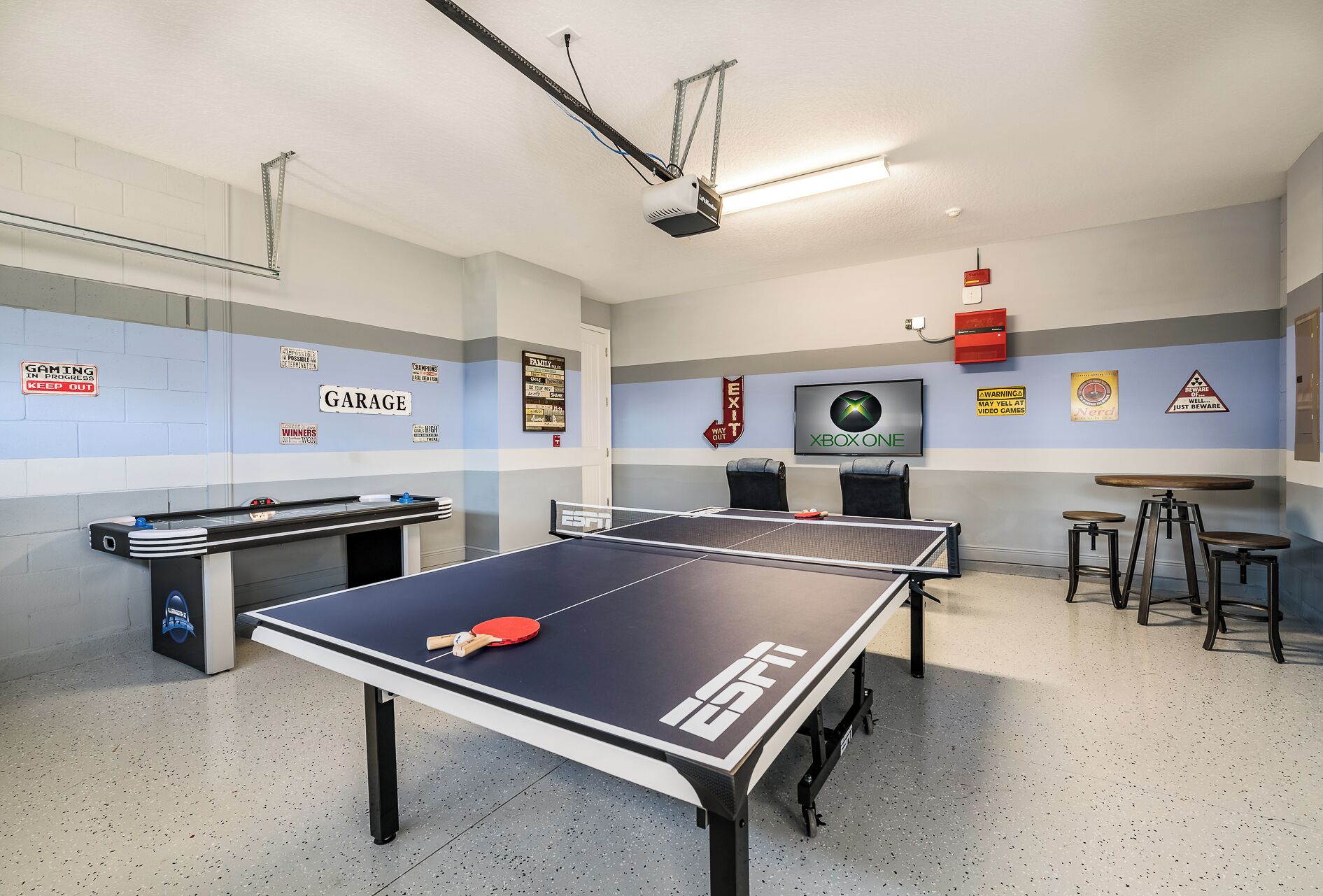 A table tennis table with a ping pong paddle and ball, a pool table, and a television screen displaying the Xbox One logo.