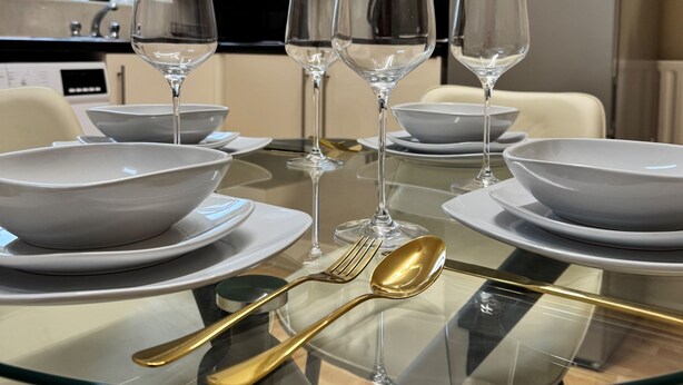 A dining table set with white plates, bowls, and gold cutlery, with three wine glasses in the center. - London City Apartment - Modern & Cozy Stay (London)