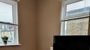 A modern living room with a flat-screen TV on a white stand, a black round table with a remote, a white sofa, and a small black stool. - London City Apartment - Modern & Cozy Stay (London)