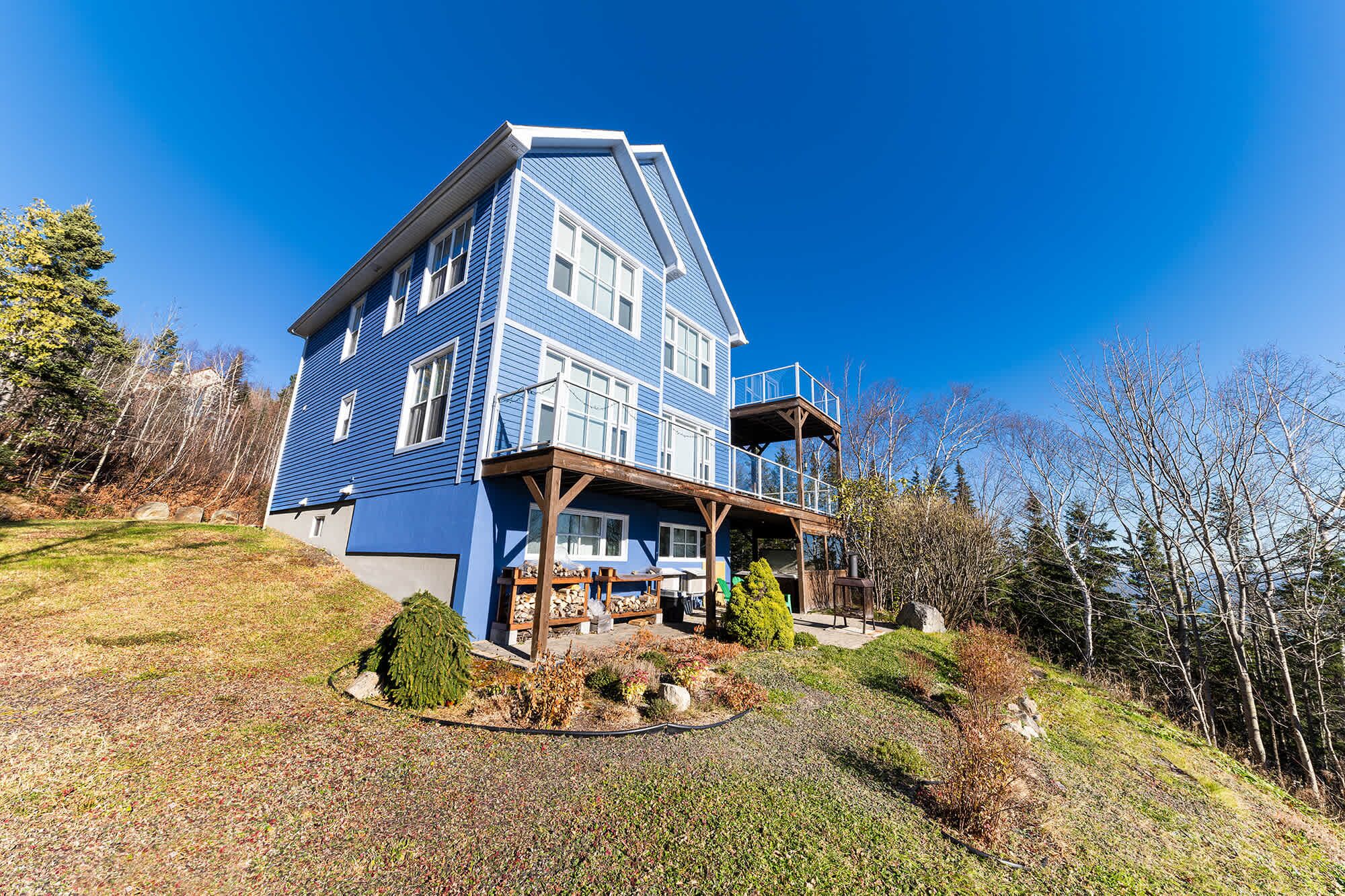 Une maison à deux étages avec une façade bleue, une terrasse en bois et un petit jardin.