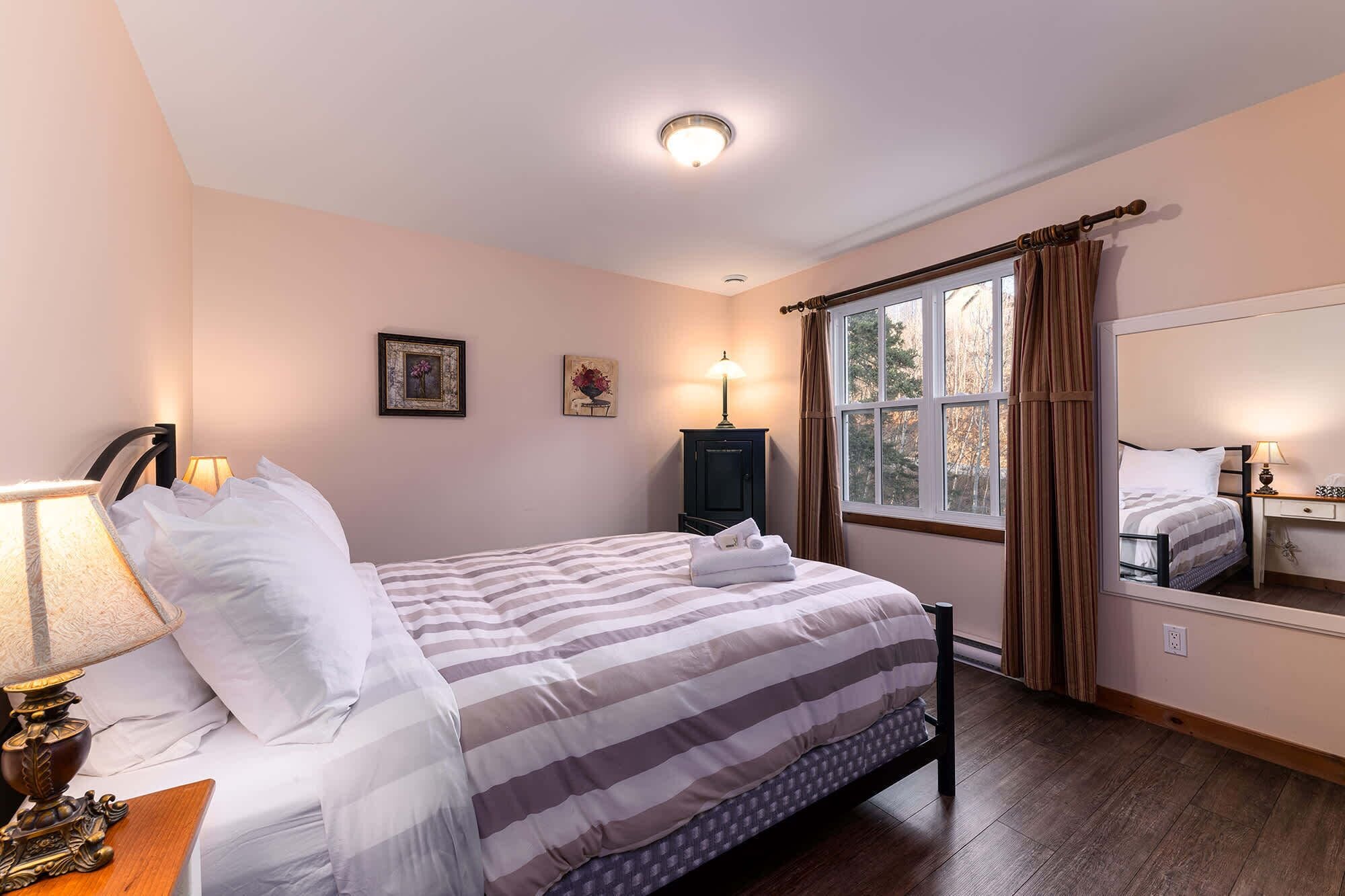 Une chambre à coucher avec un lit, deux lampes de chevet, une commode, un miroir et une fenêtre avec des rideaux.