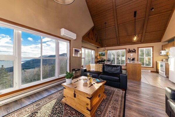 A spacious living room with large windows, a black leather sofa, a wooden coffee table, and a kitchen area with a microwave and stove. - SPA & Mountain View | Condo Lapaix-Ski (Petite-Rivière-Saint-François)