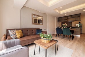 Ein modernes Wohnzimmer mit einem braunen Sofa, einem kleinen Tisch mit einem Korb und einem Essbereich mit Stühlen. Im Hintergrund ist eine Küche zu sehen.