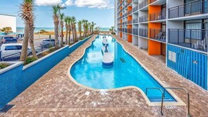 Una piscina con vista despejada al océano, rodeada de palmeras y un edificio de varios pisos con balcones.