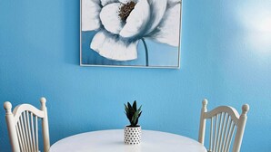 A round white table with two white chairs, a small potted plant, and a framed floral artwork on a blue wall. - SM50 Bright Studio walk to the beach (Myrtle Beach)