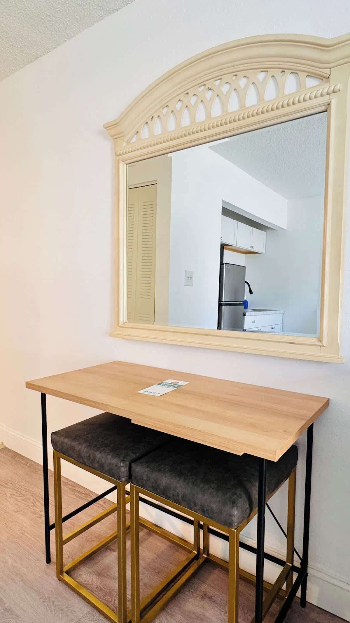 A small, square wooden desk with a black cushioned bench, a gold frame, and a mirror above it.