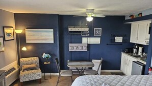 A compact hotel room with a bed, a small table, a chair, a desk, a ceiling fan, a wall-mounted shelf, and a kitchenette. - Blue Water Resort #19 great studio on the beach! (Myrtle Beach)