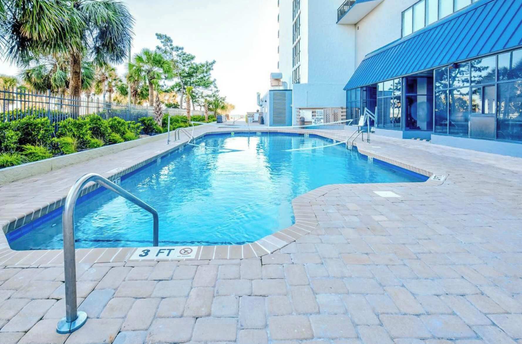 A swimming pool with a 3 FT depth marker, surrounded by a paved area with a metal railing.