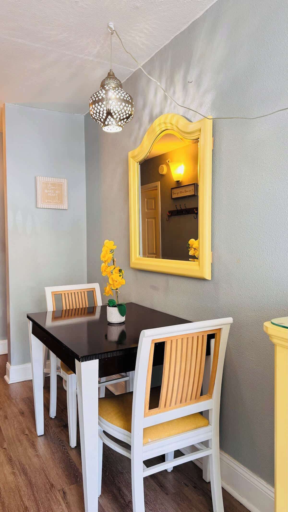 A dining area with a dark wooden table, white chairs, a yellow-framed mirror, and a hanging light fixture.