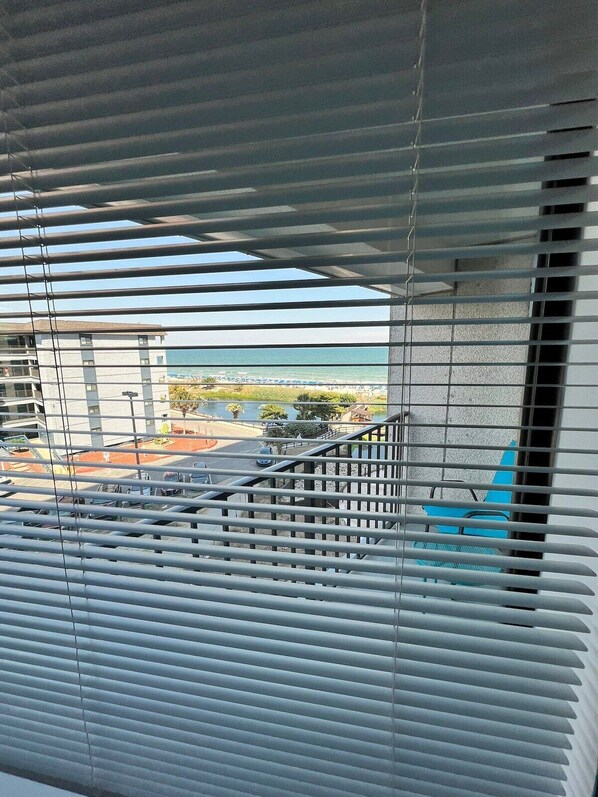 A view through window blinds, featuring a building with a balcony and a clear view of the beach and ocean.