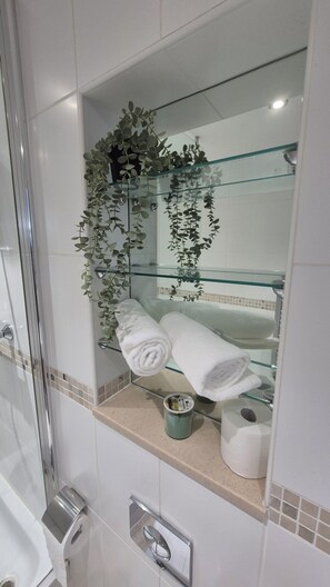 Ein Badezimmer mit einer Duschkabine aus Glas, einer Glasablage mit Handtüchern, einem Toilettenpapierhalter und einer kleinen Pflanze.