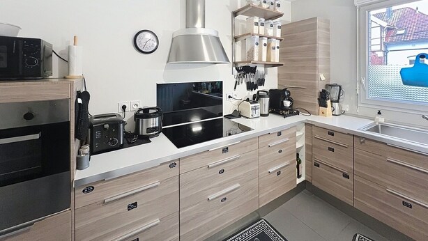 A modern kitchen with a stove, microwave, and coffee maker. There is a clock on the wall and a window with a view of a house. - Charming, cosy room just 5 minutes from Lille (Marcq-en-Barœul)